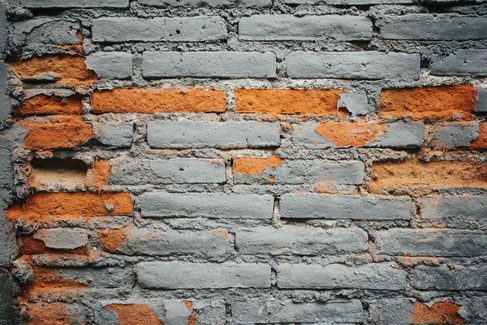 A brick wall with grey paint flaking away from orange brick and crumbling mortar joints